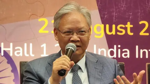 Xu Feihong / Twitter O embaixador chinês na Índia, Xu Feihong, vestindo uma camisa branca e uma jaqueta cinza e uma gravata preta e branca, falou com um microfone em um evento em Delhi na quinta-feira